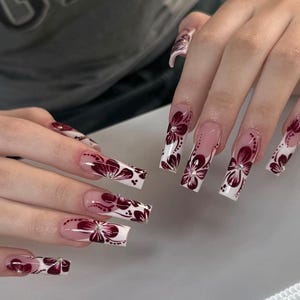 以下が含まれることがあります： クリアベースと白いチップが付いた、長くて四角いアクリルネイル。各爪には、小さなラインストーンのアクセントが付いた濃い赤色の花の模様があります。爪はよく手入れされており、詳細なネイルアートが施されています。