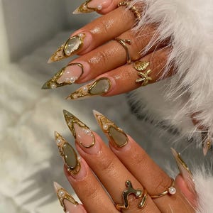 Puede incluir: Uñas largas en forma de estilete con base nude, decoradas con remolinos metálicos dorados y pequeñas perlas blancas. Los dedos lucen varios anillos dorados, incluyendo diseños con forma de concha y estrella de mar, sobre piel sintética blanca.