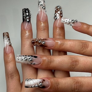 May include: Long, square-shaped artificial nails with a variety of designs. Some nails feature a black and white spotted pattern, while others have a brown and white spotted pattern. All nails are adorned with silver studs.