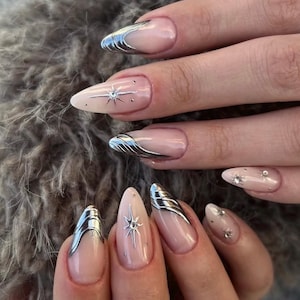 Puede incluir: Uñas en forma de almendra con una base color nude y detalles en plata metálica. El diseño incluye puntas francesas con acabado cromado y líneas onduladas, además de pequeños adornos en forma de estrella con tachuelas plateadas.