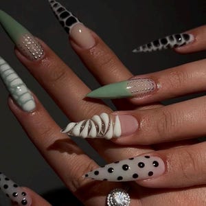 May include: A close-up of a hand with long, pointed acrylic nails. The nails feature various designs, including mint green tips with silver studs, black and white cow print, and white swirls. One nail has black polka dots.