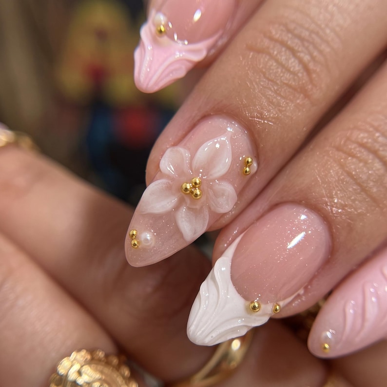 May include: Almond-shaped nails featuring a sheer nude base with a subtle shimmer. The design includes three-dimensional white floral accents, raised white sculpted tips, and small gold metallic beads and tiny white pearls as decorative embellishments.