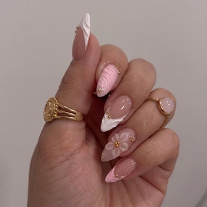 May include: Almond-shaped nails featuring a mix of white and pink designs, including 3D floral accents, textured swirls, and small gold beads. Two gold rings are worn, one with a circular embossed detail and the other with a smooth pink stone.