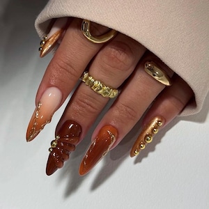 Pode incluir: Uma mão com unhas longas em formato amêndoa em tons de castanho, laranja e dourado. Algumas unhas apresentam detalhes dourados, enquanto outras têm elementos 3D. Vários anéis dourados adornam os dedos, complementando a nail art.