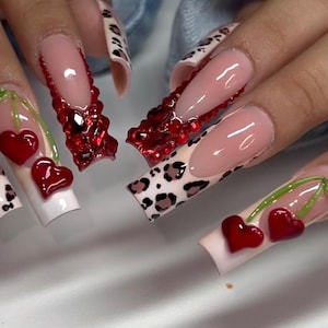 May include: A set of long, artificial nails with a variety of designs. Some nails feature a French tip with a leopard print, while others have a clear base with red heart-shaped cherries. One nail has a red jeweled design.