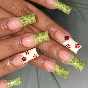 Puede incluir: Uñas artificiales largas y cuadradas. Algunas uñas presentan un diseño de cachemira verde claro, mientras que otras tienen una base blanca con detalles de flores rojas y una mariquita. Las uñas tienen una base rosa y una punta francesa.