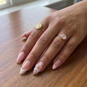 May include: Two gold rings worn on fingers. One ring features a circular medallion with a raised figure, while the other holds a smooth, round, pale pink stone. The fingernails are shaped in a long almond style with light pink and white floral designs.