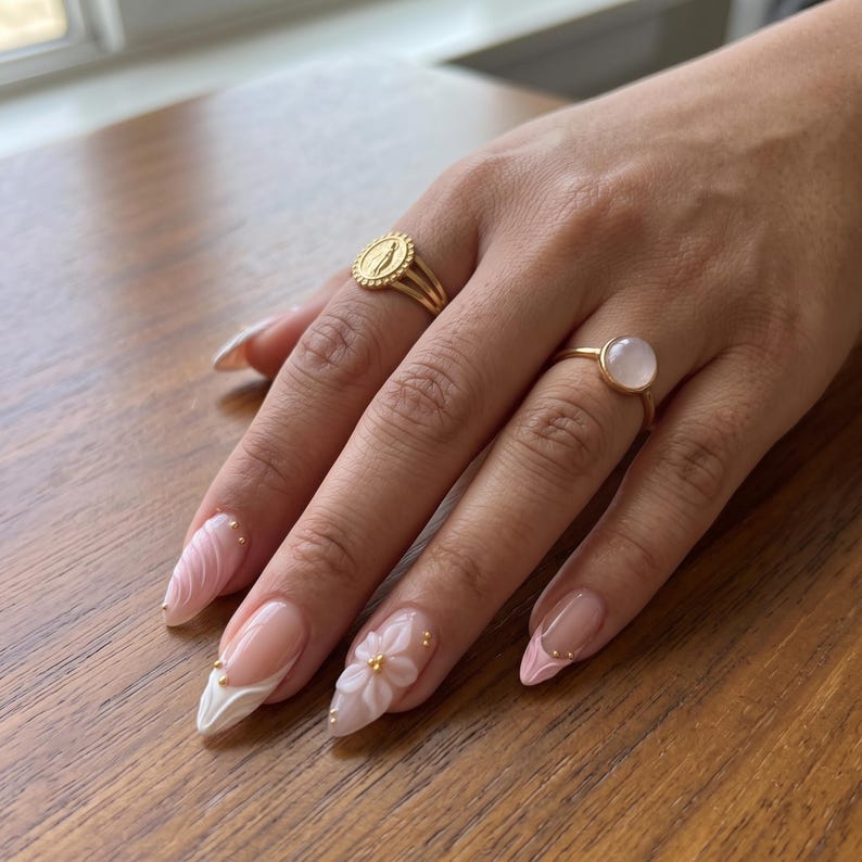 May include: Two gold rings worn on fingers. One ring features a circular medallion with a raised figure, while the other holds a smooth, round, pale pink stone. The fingernails are shaped in a long almond style with light pink and white floral designs.