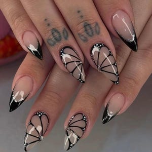 Könnte beinhalten: Nahaufnahme einer Hand mit langen, spitzen Nägeln, die mit einem schwarz-weißen Schmetterlingsdesign bemalt sind. Die Nägel sind außerdem mit einer dünnen Linie aus silbernem Glitzer verziert.