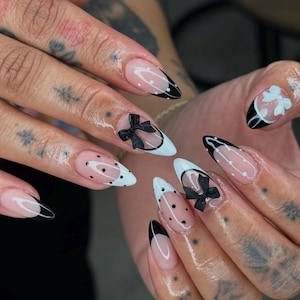 May include: Close-up of a hand with long, almond-shaped nails painted with a clear base and black and white accents. Some nails feature black polka dots, while others have black bows. The nails are styled with a white tip and a black outline.