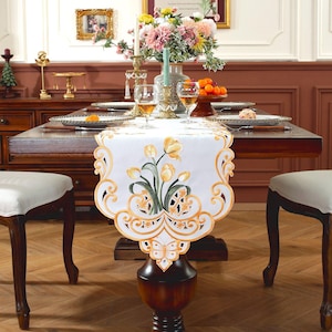Könnte beinhalten: Ein weißer Tischläufer mit gestickten gelben Tulpen und einem Schmetterlingsmuster. Der Läufer liegt auf einem Holztisch mit zwei Stühlen.