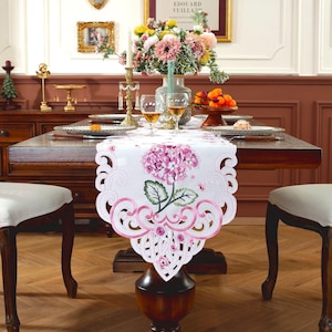 Könnte beinhalten: Weißer Tischläufer mit einem rosa gestickten Hortensien-Design. Der Läufer hat einen gewellten Rand mit rosa Akzenten. Der Tisch ist mit Tellern, Gläsern und einem Blumengesteck gedeckt, wodurch eine elegante Essumgebung entsteht.