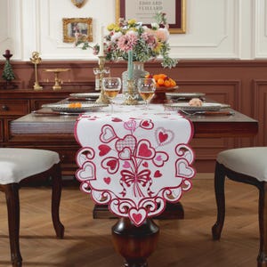 May include: White table runner with a red and pink embroidered heart design. The runner features a bouquet of hearts with a butterfly in the center. The runner is on a wooden table with two chairs.