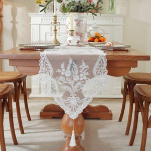 Könnte beinhalten: Weißer Tischläufer aus Spitze mit floralem Muster und Quastenfransen. Der Läufer liegt auf einem Holztisch mit zwei Stühlen.