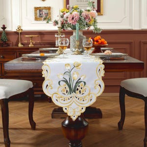 Könnte beinhalten: Weißer Tischläufer mit gestickten gelben Tulpen und grünen Blättern. Der Läufer hat einen gewellten Rand mit einem goldenen Spitzenmuster.