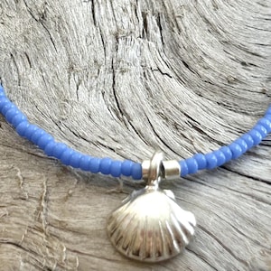 May include: A blue beaded necklace with a silver seashell pendant. The necklace is made of small, round, light blue beads. The pendant is a detailed, silver seashell. The necklace is displayed on a weathered, gray wooden surface.