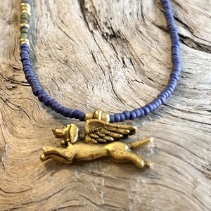 May include: A necklace featuring a brass pendant of a dog with wings, suspended from a beaded blue necklace. The necklace has a rustic, antique appearance. The beads are a deep blue color, and the pendant is a warm gold tone.