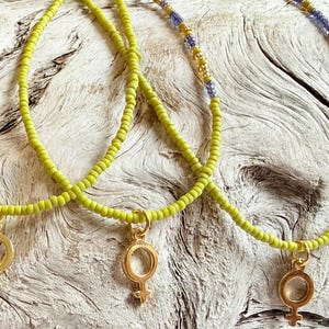 May include: Three beaded necklaces with a gold-colored female symbol pendant. The necklaces feature small, light green beads with sections of gold and blue beads. The necklaces are displayed on a light-colored, textured wooden surface.