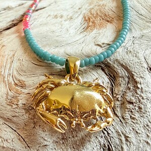 May include: A gold-colored crab pendant necklace with a turquoise beaded chain. The necklace has a lobster clasp. The beads transition to pink and gray near the clasp. The necklace is displayed on a weathered wood surface.
