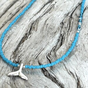 May include: A turquoise beaded necklace with a silver whale tail pendant. The necklace is made of small, round beads and is displayed on a weathered wood surface. The pendant is a detailed, stylized whale tail.