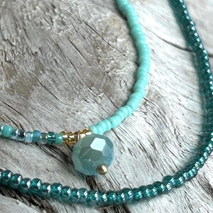 May include: Close-up of two beaded necklaces. One necklace is turquoise with a faceted blue-green pendant and gold accents. The other necklace is a darker teal color. The necklaces are displayed on a weathered wood surface.
