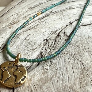 May include: A beaded necklace with a gold-colored pendant featuring a world map design. The necklace has a teal and green color scheme, with small beads strung together. The pendant is round and has a detailed depiction of the continents.