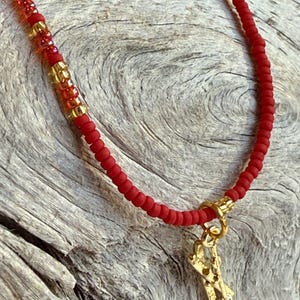 May include: Red beaded necklace with gold accents and a gold-colored charm. The necklace features small red beads and larger gold beads. The charm is a gold-colored diving equipment pendant.