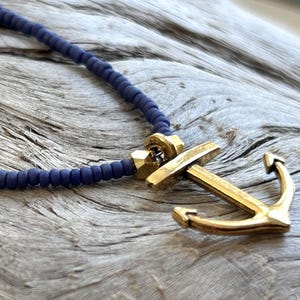 May include: A nautical-themed necklace featuring a gold-colored anchor pendant and a dark blue beaded chain. The anchor is detailed with a classic design. The necklace rests on a weathered, gray wooden surface.