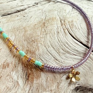 May include: A beaded necklace with a gold flower pendant. The necklace features a combination of light blue, gold, and purple beads. The flower pendant is a simple, five-petal design. The necklace is displayed on a light-colored, textured wooden surface.