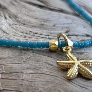 May include: A turquoise beaded necklace with a gold starfish pendant. The necklace is made of small, round beads and has a gold clasp. The starfish pendant is detailed and textured, adding a touch of coastal charm.