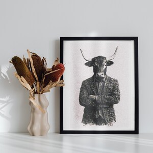 Könnte beinhalten: Schwarz gerahmter Kunstdruck mit einem Stier mit menschlichem Körper, der eine Tweedjacke trägt. Links vom Druck befindet sich eine beige Vase mit getrockneten Blumen. Der Kunstdruck und die Vase stehen auf einer weißen Oberfläche.