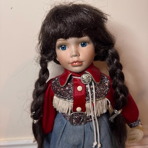 May include: A porcelain doll with blue eyes and dark brown braided hair. The doll wears a red velvet jacket with fringe and a denim skirt. A silver buckle and decorative buttons adorn the jacket. The doll has a realistic face with painted features.