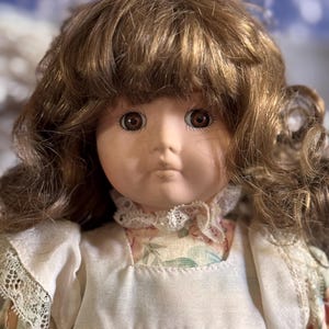 May include: A vintage porcelain doll with brown hair and eyes. The doll is dressed in a cream-coloured dress with a floral pattern and lace trim around the collar and sleeves. The doll's hair is styled in loose curls.