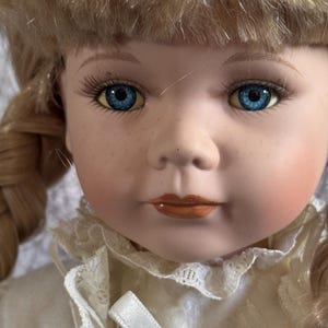 May include: A porcelain doll with blonde hair styled in braids, bright blue eyes, long eyelashes, and light freckles across the nose. The doll features a pale complexion with rosy cheeks and painted orange lips, wearing a cream-coloured lace collar.