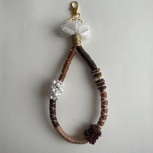 Op de afbeelding: Een druppelvormige sleutelhanger omwikkeld met bruin en beige garen, voorzien van een witte kwast aan de bovenkant, een gouden karabijnhaak, een cluster witte kunstparels, een sectie met veelkleurige kraaltjes en een donkerrode kralenbloem.