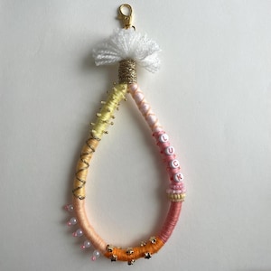 Op de afbeelding: Een druppelvormige sleutelhanger omwikkeld met meerkleurig garen, voorzien van een witte kwast aan de bovenkant met een gouden karabijnsluiting. Het garen verloopt van geel naar roze en oranje, versierd met kleine kraaltjes, sterretjes en witte letterkralen die LUCK spellen.