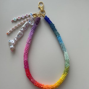 Op de afbeelding: Een kralen sleutelhanger met een regenboogkleurverloop. De sleutelhanger heeft een goudkleurige sluiting en een bedel met het woord "LUCKY" in gekleurde letterkralen. Extra bedels zijn witte dobbelsteen-vormige kralen.