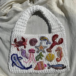 Puede incluir: Un bolso tote de crochet blanco adornado con criaturas marinas de cuentas de colores. El bolso presenta un cangrejo, una langosta, un caballito de mar, una estrella de mar, un pez y otra vida marina. El bolso tiene un asa gruesa y texturizada.