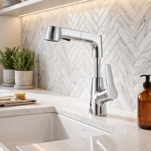 Può includere: Un rubinetto da cucina in cromo lucido con doccetta estraibile e una singola leva bianca, montato su un piano di lavoro in marmo bianco accanto a un lavello in ceramica bianco, un panno beige, una spazzola in legno e un dispenser di sapone ambrato.