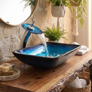 May include: A rectangular glass vessel sink with a deep blue and black gradient finish, featuring a matching blue glass waterfall faucet. The sink is mounted on a natural live-edge wooden vanity top, with water flowing from the faucet into the basin.
