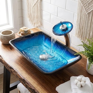 Könnte beinhalten: Ein rechteckiges Aufsatzwaschbecken aus Glas mit einem leuchtend blauen Farbverlauf. Ein Wasserfall-Wasserhahn aus Chrom mit einer passenden blauen Glasscheibe oben lässt Wasser in das Becken fließen. Das Waschbecken hat einen polierten Chrom-Ablauf.