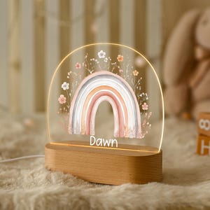 May include: A rainbow-shaped night light with a wooden base. The design features pastel colours and floral details. The name "Dawn" is printed on the light. The light is on, illuminating the rainbow design.