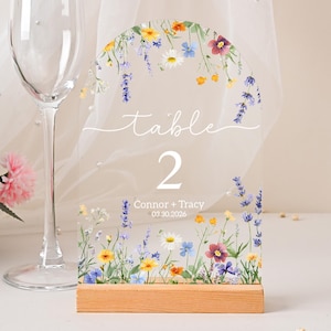 May include: Clear acrylic table number with floral design. The number "2" is displayed, along with the word "table" in script. The base is made of wood, and the design includes colorful flowers. The names "Connor + Tracy" and the date "03.30.2026" are also visible.