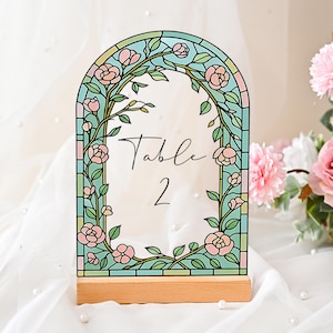 May include: A decorative table number in a stained-glass effect, featuring a floral design. The arched frame is adorned with pink roses and green foliage, with the text "Table 2". It is set on a wooden base, suitable for weddings or special events.