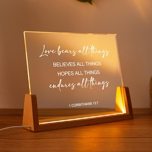 May include: A lighted acrylic sign with a wooden base. The sign displays the text "Love bears all things, believes all things, hopes all things, endures all things" and "1 Corinthians 13:7" in a white script font. The base is made of light-colored wood.