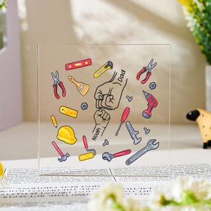 Puede incluir: Una placa cuadrada de acrílico transparente con una ilustración colorida de varias herramientas de construcción, incluyendo un casco amarillo, alicates rojos, una llave inglesa y un taladro. En el centro, un gráfico de puños muestra los nombres Dad y Noah.