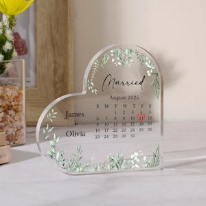 Può includere: Calendario di nozze a forma di cuore in acrilico trasparente con la parola "Married" e il mese di agosto 2025. I nomi James e Olivia sono elencati, con l'11 agosto evidenziato in rosso. Accenti floreali decorano i bordi.