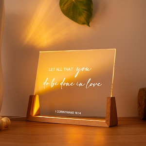 Puede incluir: Un letrero acrílico iluminado con el texto "LET ALL THAT you do be done in love" y "1 CORINTIOS 16:14" en escritura blanca. El letrero está colocado en una base de madera, iluminado con un brillo cálido, y es un artículo decorativo.