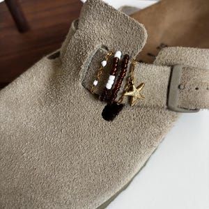 Op de afbeelding: Een taupekleurige suède klomp met een decoratieve band versierd met drie kralenbedels. De bedels bestaan uit een goudkleurige ketting met witte kralen, een streng donkerbruine kralen en een goudkleurige zeesterhanger aan een metalen ring.