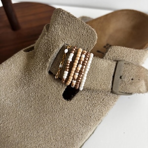 Op de afbeelding: Een taupekleurige suède klomp met een decoratieve band versierd met een goudkleurige ketting en een reeks kleine, veelkleurige kralen in tinten wit, crème en metallic brons, vastgezet met een bijpassende taupekleurige gesp aan de zijkant.